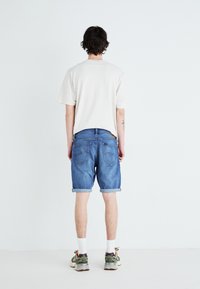 Camiseta beige de cuello redondo, pantalones cortos de denim con el dobladillo enrollado, color azul, vista trasera. El modelo lleva calcetines blancos y zapatillas verdes/grises.