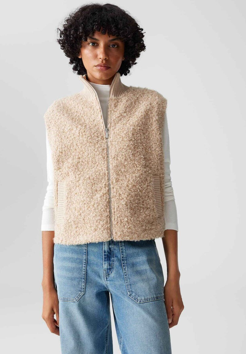 Gilet in pile beige con colletto alto, zip frontale e tasche laterali, abbinato a una maglietta bianca a maniche lunghe e jeans azzurri chiari.