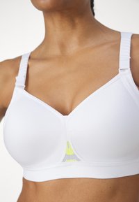 Witte sportbh met een V-vormig design, gestructureerde stof, verstelbare schouderbanden en een mesh paneel met een gele accent in het midden.