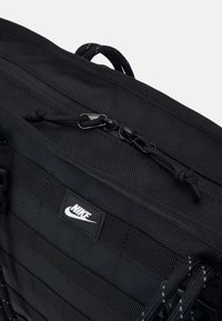 Must be tekstureeritud nailonist must seljakott. Omab lukuga ülemist osa, reguleeritavaid rihmasid ja silmatorkavat valget Nike logo kangapatchil.