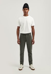Witte kortegalimde t-shirt, olijfgroene broek, zwarte beanie, en witte sneakers met bruine zolen, gepresenteerd tegen een neutrale achtergrond.