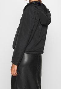 Svart puffjacka med huva, med en fuskpälsfoder längs kragen. Den har en kort klippning och tryckknappsdetaljer på ärmarna.