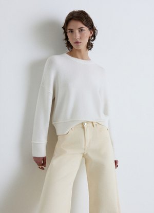 Young woman with short brown hair wears a loose white sweater and high-waisted wide-leg cream pants against a plain white background.
