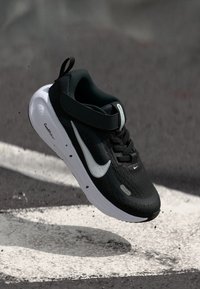 Schwarzer Sportschuh mit einem strukturierten Obermaterial, weißem Swoosh-Logo, verstellbarem Riemen und weißer Dämpfungsohle. Schwebt über einer strukturierten Fläche.