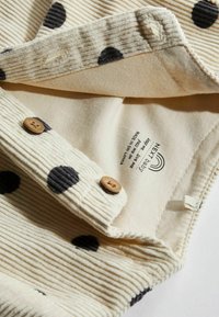 Beige corduroy stof met grote zwarte stippen, met twee houten knopen en een label met de tekst "NEXT Baby, gemaakt in Sri Lanka."