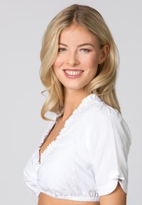 Junge Frau mit blonden Haaren, die lächelt und eine weiße Rüschentbluse vor einem hellgrauen Hintergrund trägt.