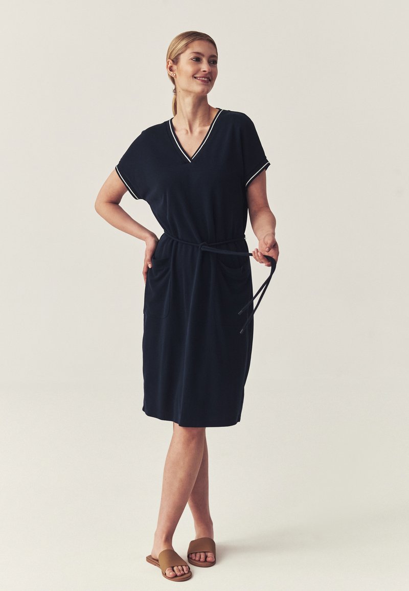 Femme debout dans une robe noire mi-longue avec un bord blanc, tenant une ceinture, portant des sandales plates marron sur un fond uni.