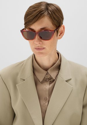 Persona con capelli corti castani che indossa occhiali da sole rettangolari marroni, giacca da completo beige e camicia taupe abbottonata su uno sfondo semplice.