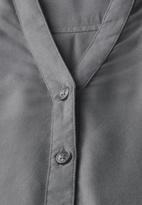 Chemise grise à manches longues avec une texture de tissu lisse, un col pointu et des boutons en plastique sombre. Design simple avec des lignes épurées.