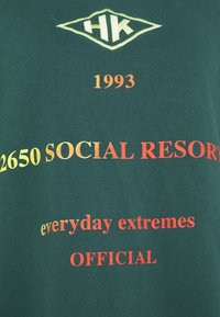 Zelené bavlněné tričko s potiskem; obsahuje logo "HK", rok "1993" a fráze v žluté, oranžové a červené barvě. Standardní střih.