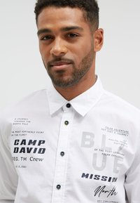 White button-up shirt featuring black and gray text graphics, a spread collar, and black buttons with green accents. Text includes brand and theme details.