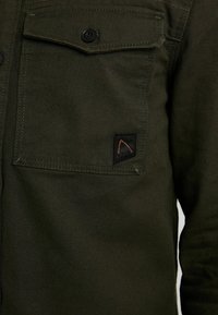 Veste en tissu vert foncé avec une poche poitrine boutonnée ornée d'un petit patch logo triangulaire noir et orange.