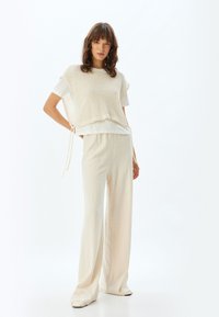Lichtgewicht crèmekleurige gebreide trui over een witte top, gecombineerd met crèmekleurige wijde broek met textuur; met trekkoordaccenten.