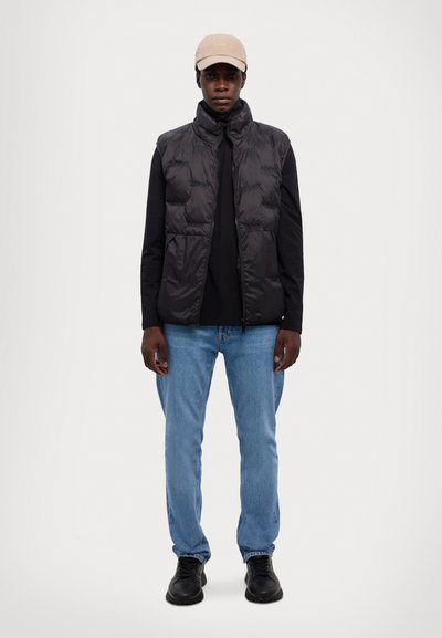 Μαύρη φούτερ γιλέκο πάνω από μαύρο μακρυμάνικο πουκάμισο, με ανοιχτό μπλε τζιν και μαύρα παπούτσια. Μπέζ καπέλο ολοκληρώνει την εμφάνιση. Λείος υφασμάτινος χαρακτήρας.