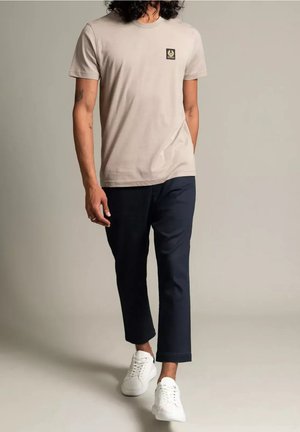 Mand med krøllet hår iført beige t-shirt, sorte korte bukser og hvide sneakers, der går foran en ensfarvet lys baggrund.