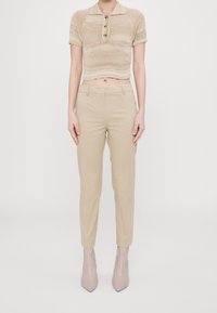 Béžové zkrácené pletené polo tričko s knoflíky, kombinované s béžovými kalhotami na míru a světle růžovými kotníkovými botami s špičatou špičkou, na neutrálním pozadí.