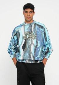 Multifärgad sweatshirt med en blandning av blå, svart och vita abstrakta mönster. Ribbad halsringning och manschetter med grafisk text på framsidan.