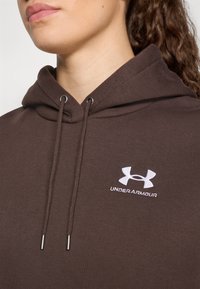 Brun hettegenser med snøring i hetten, med sølvfargede metallringer og en hvit brodert Under Armour-logo på brystet.