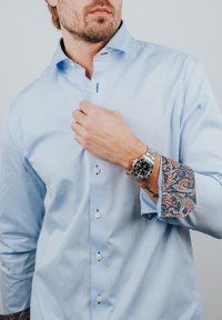 Lichtblauw overhemd met een patroon op de manchetten. Heeft een klassieke kraag, zes knopen en een contrasterend paisley-design op de manchetten.