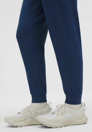Witte en beige sportchoenen met textuurelementen, gecombineerd met marineblauwe joggingbroek met elastische boorden. Zachte gebreide sokken zichtbaar boven de schoenen.