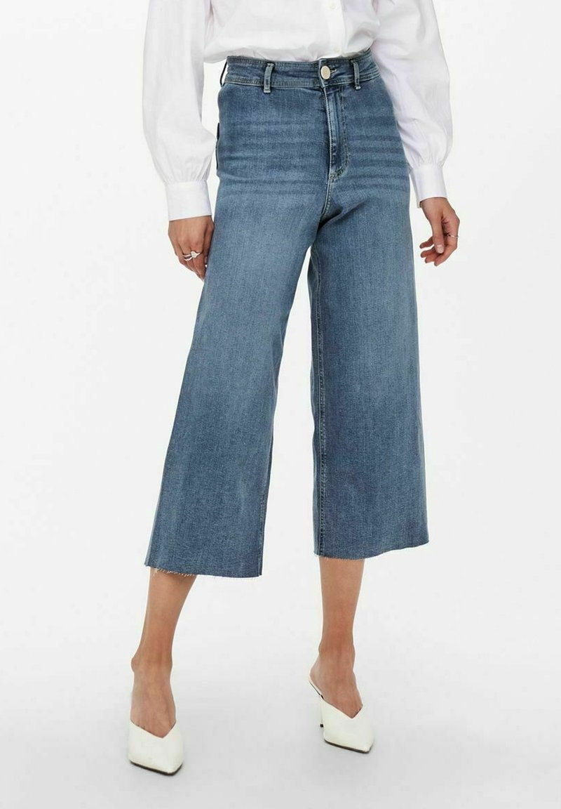 Personne portant un jean bleu taille haute, jambes larges et raccourci, un chemisier blanc à manches longues, et des chaussures blanches à talons pointus.