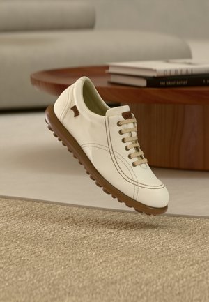 Chaussure en cuir blanc avec semelle marron, coutures décoratives et design à lacets. Dispose d'un petit logo marron sur le côté. Flottant au-dessus d'une surface texturée.