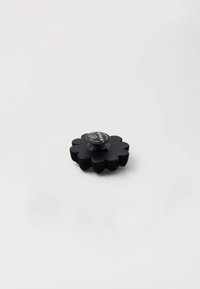Pulsante nero a forma floreale con superficie testurizzata e protuberanza centrale, realizzato in un materiale morbido. Logo visibile in alto.