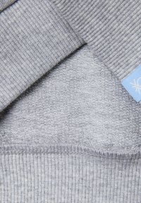 Primer plano de tela gris claro doblada que muestra texturas acanaladas y con bucles, con una pequeña etiqueta azul parcialmente visible.