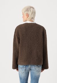 Veste en polaire brun, texturée, à coupe ample et à manches longues. Dotée d'un col rond et sans fermetures visibles. Associée à un jean clair.