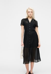 Ξανθιά γυναίκα που φοράει μαύρο midi φόρεμα με κουμπιά, κρατώντας μια λευκή τσάντα, στέκεται μπροστά σε απλό λευκό φόντο.