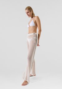 White bikini top with thin straps paired with sheer, open-knit, long white pants featuring black side seams. Model stands barefoot, gaze downward.