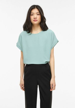 Femeie cu păr negru până la umeri, purtând o bluză largă cu mâneci scurte de culoare verde deschis și pantaloni negri cu talie înaltă, stând pe fundal alb.