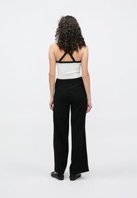 Pantalon noir à jambes larges, taille élastique, associé à un haut blanc ajusté avec des bretelles noires. Cheveux bouclés visibles de dos.