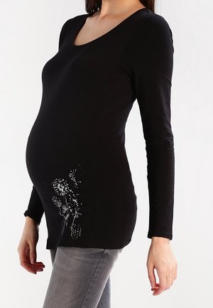 Schwangere Frau trägt ein langärmliges schwarzes Oberteil mit weißem Blumenmuster und graue Jeans, posiert vor einem weißen Hintergrund.