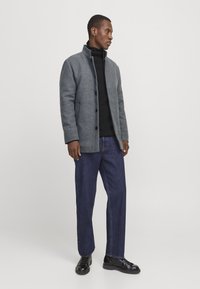 Homme portant un manteau gris boutonné, un col roulé noir, un jean bleu et des chaussures noires à lacets, se tenant devant un fond blanc uni.