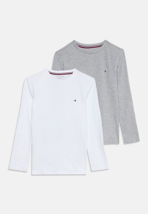 Twee lange mouwen crewneck-shirts, één wit en één grijs, elk met een klein vlaglogo op de linkerborst.