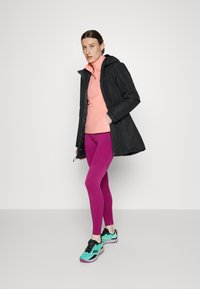 Svart vanntett parkas, rosa fleece-genser, lilla leggings og teal joggesko med svarte detaljer. Modellen står mot en enkel bakgrunn.