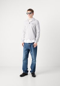 Grå zip-up genser med logo, hvit skjorte lagdelt under, blå jeans og svarte sko; modellen har på seg firkantede solbriller.