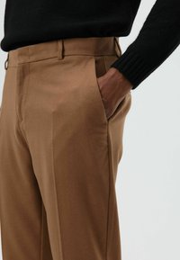 Bruine, op maat gemaakte broek van zachte stof, met zakken aan de zijkant en een strakke, slanke pasvorm met strakke lijnen. Draagt een zwarte trui.