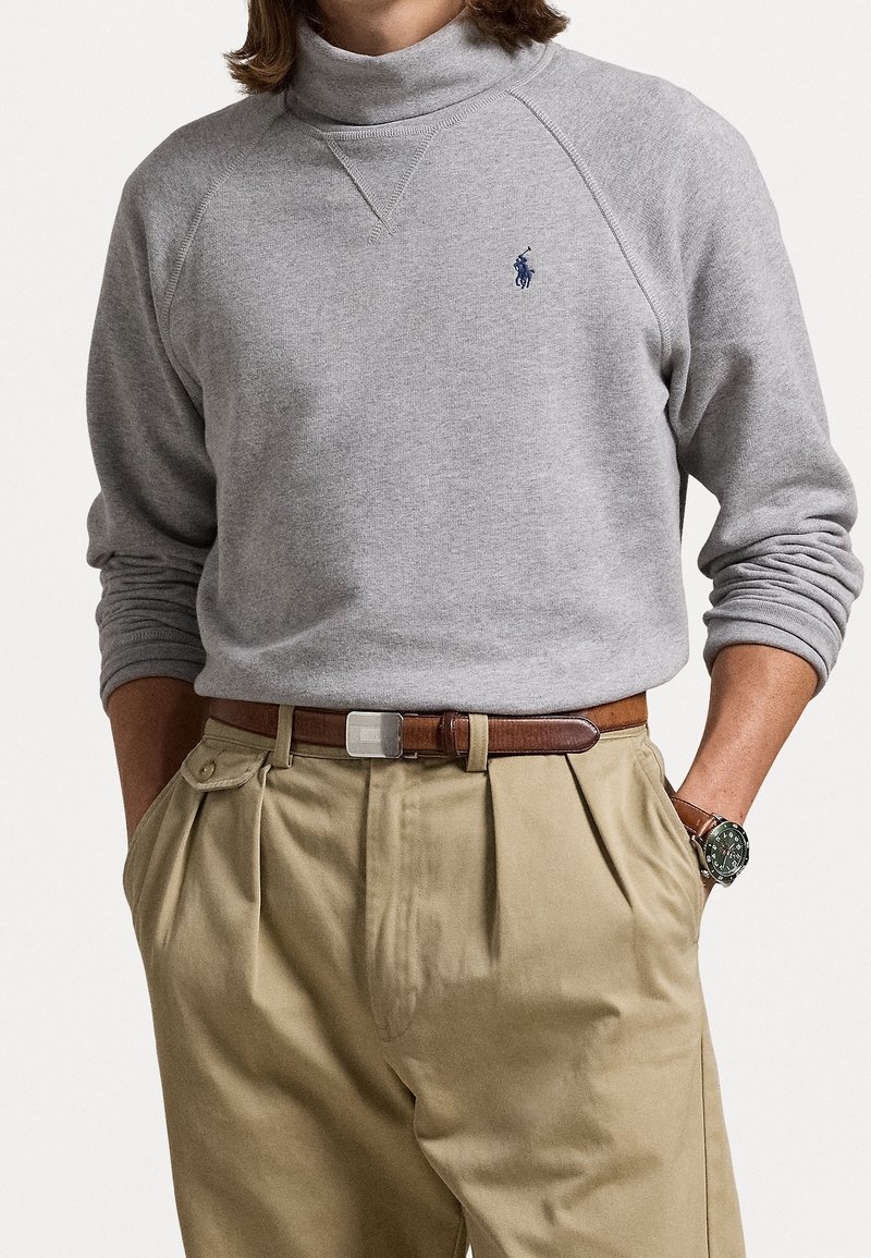 Sudadera gris de manga larga con cuello alto y un pequeño logo azul, combinada con pantalones caqui claros y un cinturón marrón. La textura parece suave.