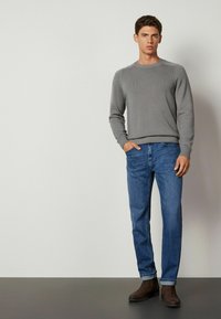 Grå stickad tröja med rund halsringning och ribbat nederkant, tillsammans med blå denimjeans. Modellen bär mörkbruna ankelboots och står på en mörk yta.