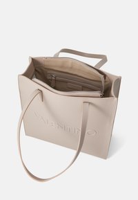 Beige tote-väska med en texturerad yta, utrustad med två långa handtag, ett zip-område och ett präglat märke på framsidan.