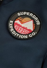 Runder aufgenähter Patch auf navyfarbigem Stoff mit Bergen, Sonne und dem Text "SUPERDRY EXPEDITION CO." in schwarz-weißem Rand.