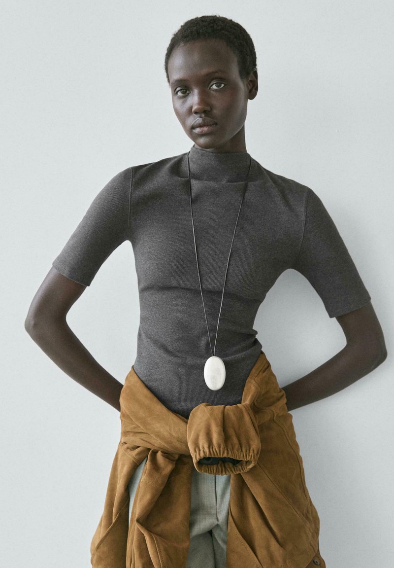 Frau mit kurzen Haaren, die ein eng anliegendes graues Oberteil, eine lange Halskette mit weißem Anhänger und eine braune Jacke, die um ihre Taille gebunden ist, trägt und vor einer schlichten Wand steht.