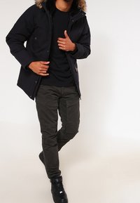 Černá parka s kapucí ozdobenou kožešinou, nošená přes černou košili a olivově zelené cargo kalhoty. Celý outfit dokonalé doplňují tenisky.