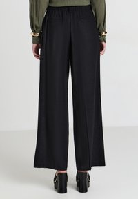 Persona che indossa pantaloni neri a gamba larga e scarpe con tacco alto nere con borchie dorate, in piedi su uno sfondo neutro.