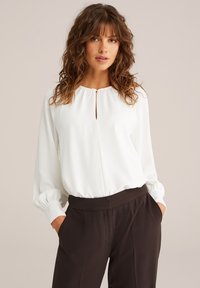 Frau mit welligem braunem Haar, die eine weiße Bluse mit langen Ärmeln und schwarze maßgeschneiderte Hose trägt, mit den Händen in den Taschen, neutraler Hintergrund.