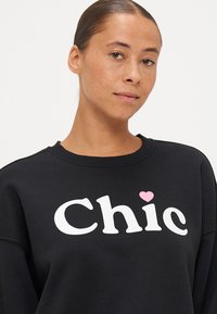 Μαύρη μπλούζα φούτερ σε χαλαρή εφαρμογή με μια μεγάλη λευκή εκτύπωση "Chic" που έχει ροζ καρδιά ως λεπτομέρεια. Μαλακό υλικό με σχέδιο στρογγυλής λαιμόκοψης.