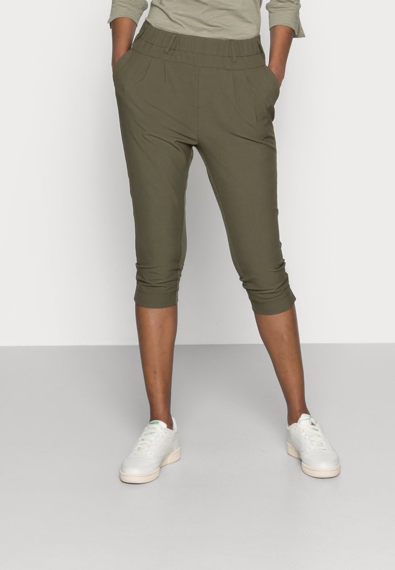 Henkilö, jolla on päällä oliivinvihreät lyhyet housut, joissa on joustava vyötärö ja valkoiset tennarit, seisoo vaaleanharmaalla lattialla valkoista seinää vasten.