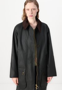 Casaco verde escuro com uma gola de bombazina castanha, grandes bolsos frontais e fecho de correr com botões. Textura lisa e design de corte reto.
